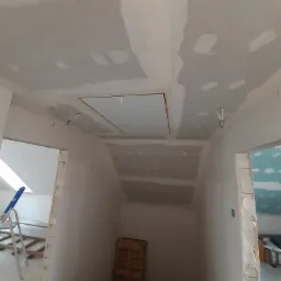 Sufit po szpachlowaniu, widoczne łączenia płyt karton-gips i właz rewizyjny, fragment instalacji elektrycznej zwisającej z sufitu, w tle fragment drabiny aluminiowej i butelki z wodą.