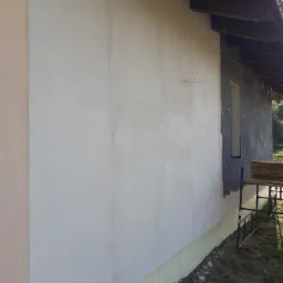 Fragment elewacji domu w trakcie prac: ocieplenie i tynkowanie, widoczne rusztowanie i niezabezpieczone okno.