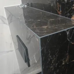 Półka z czarnego marmuru z widocznym włącznikiem światła, zamontowana w rogu łazienki wyłożonej białymi płytkami imitującymi marmur.