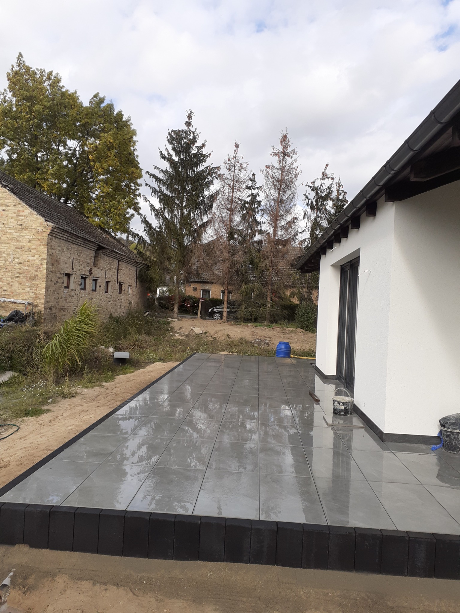 Wykończony taras z szarych płytek, otoczony czarną obwódką, przylegający do białej ściany budynku z czarnymi oknami, w tle stary ceglany budynek i drzewa.