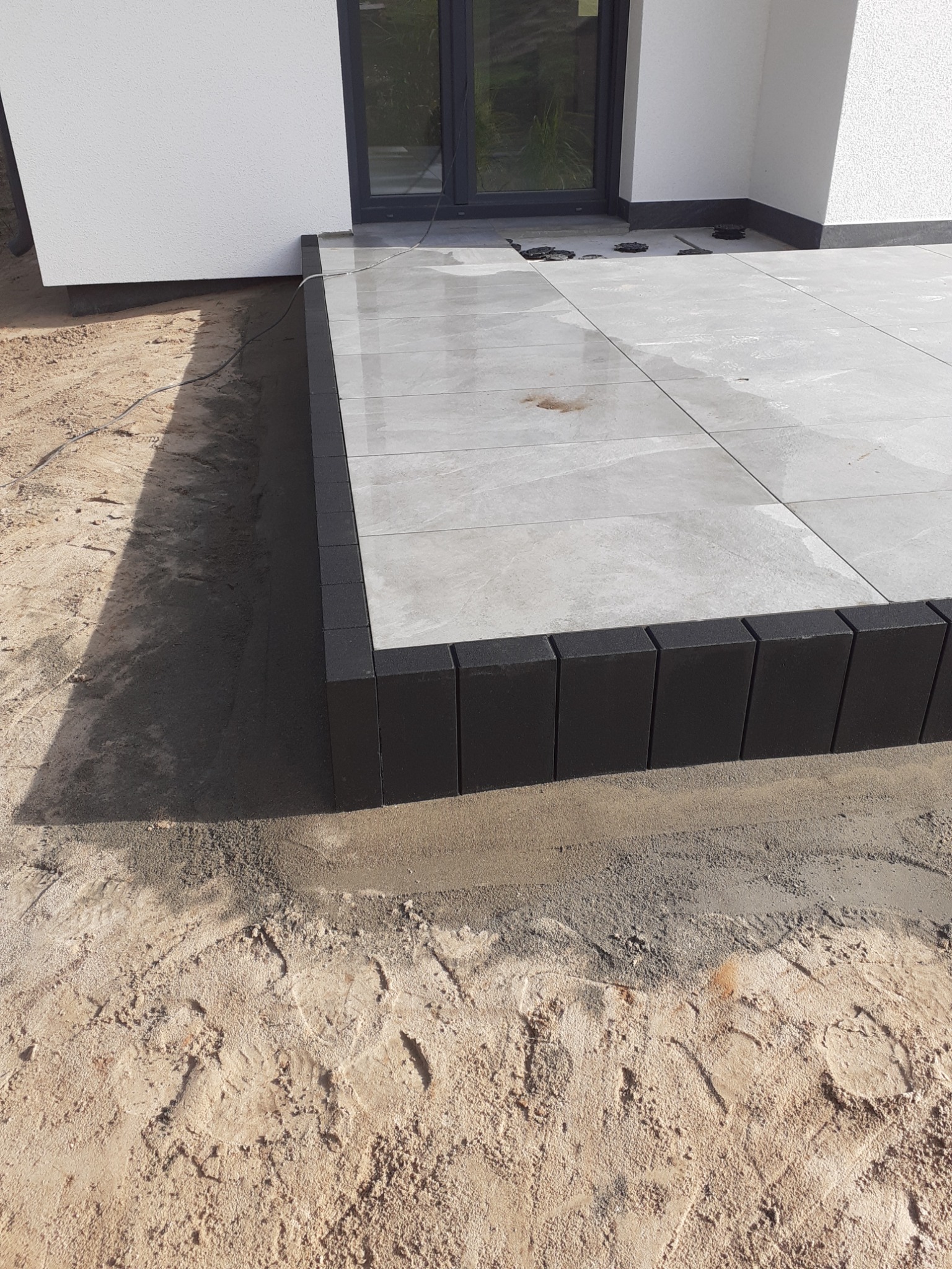 Nowy taras z szarych płytek ceramicznych obramowany czarnymi kostkami, widoczny fragment elewacji budynku z oknem tarasowym, piasek wokół.