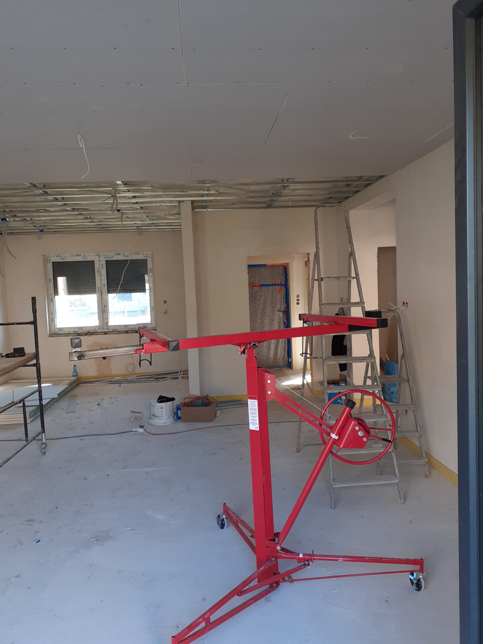 Wnętrze w trakcie montażu płyt gipsowo-kartonowych na suficie, widoczny podnośnik do płyt w kolorze czerwonym, częściowo zamontowany stelaż i drabina aluminiowa.