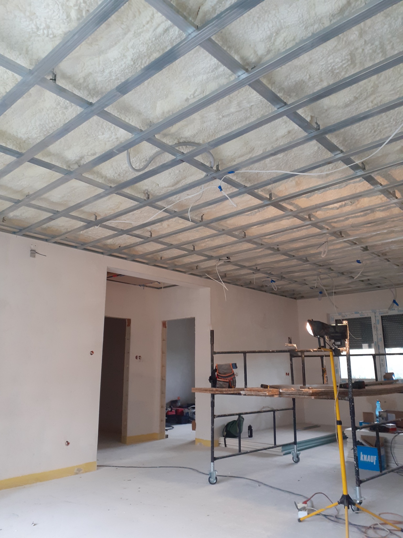 Wnętrze w trakcie prac: metalowa konstrukcja sufitu podwieszanego z widoczną warstwą izolacji z pianki natryskowej, oświetlenie budowlane na trójnogu, rusztowanie z narzędziami, przygotowane ściany...