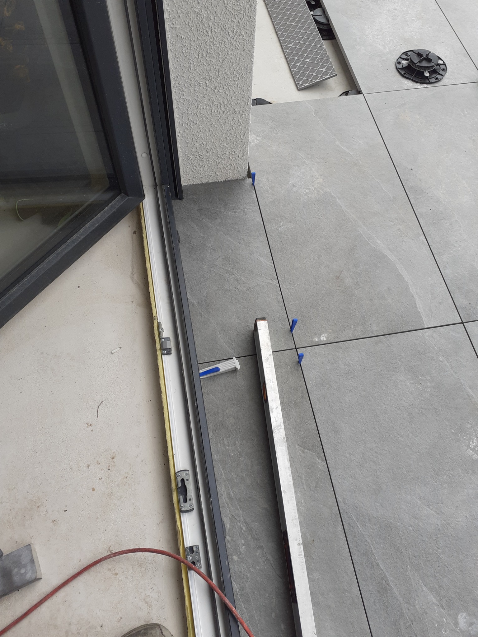 Montaż szarych płytek na balkonie, widoczny poziomica, krzyżyki dystansowe i podkładki poziomujące pod płytkami, okno balkonowe z czarną ramą.