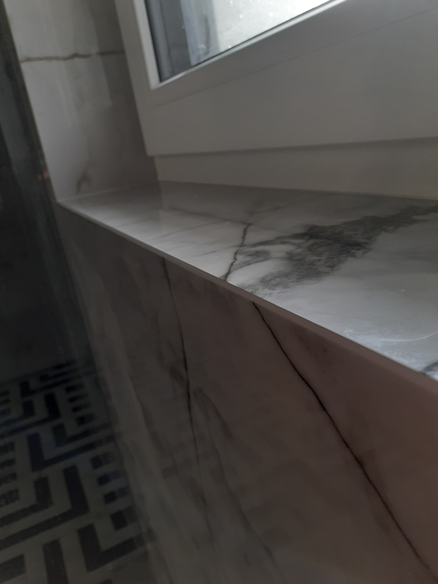 Marmurowy parapet okienny w łazience, z widoczną strukturą kamienia i białymi ramami okiennymi, oraz fragmentem podłogi z wzorem geometrycznym w tle.