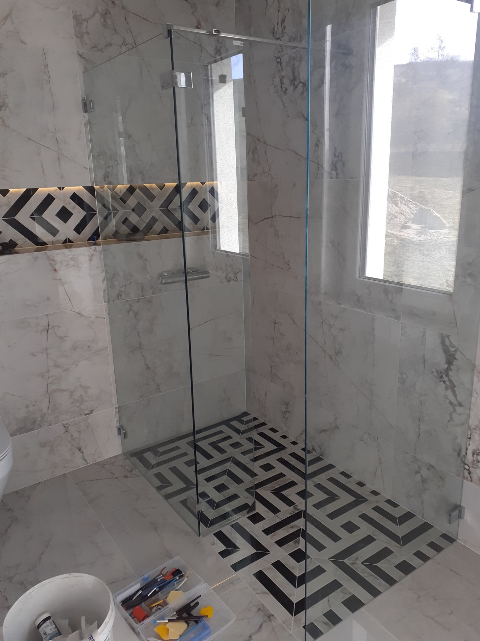 Nowoczesna kabina prysznicowa z przezroczystego szkła, wykończona marmurowymi płytkami i wzorzystą posadzką, widoczny fragment toalety oraz narzędzia w wiadrze i pojemniku.