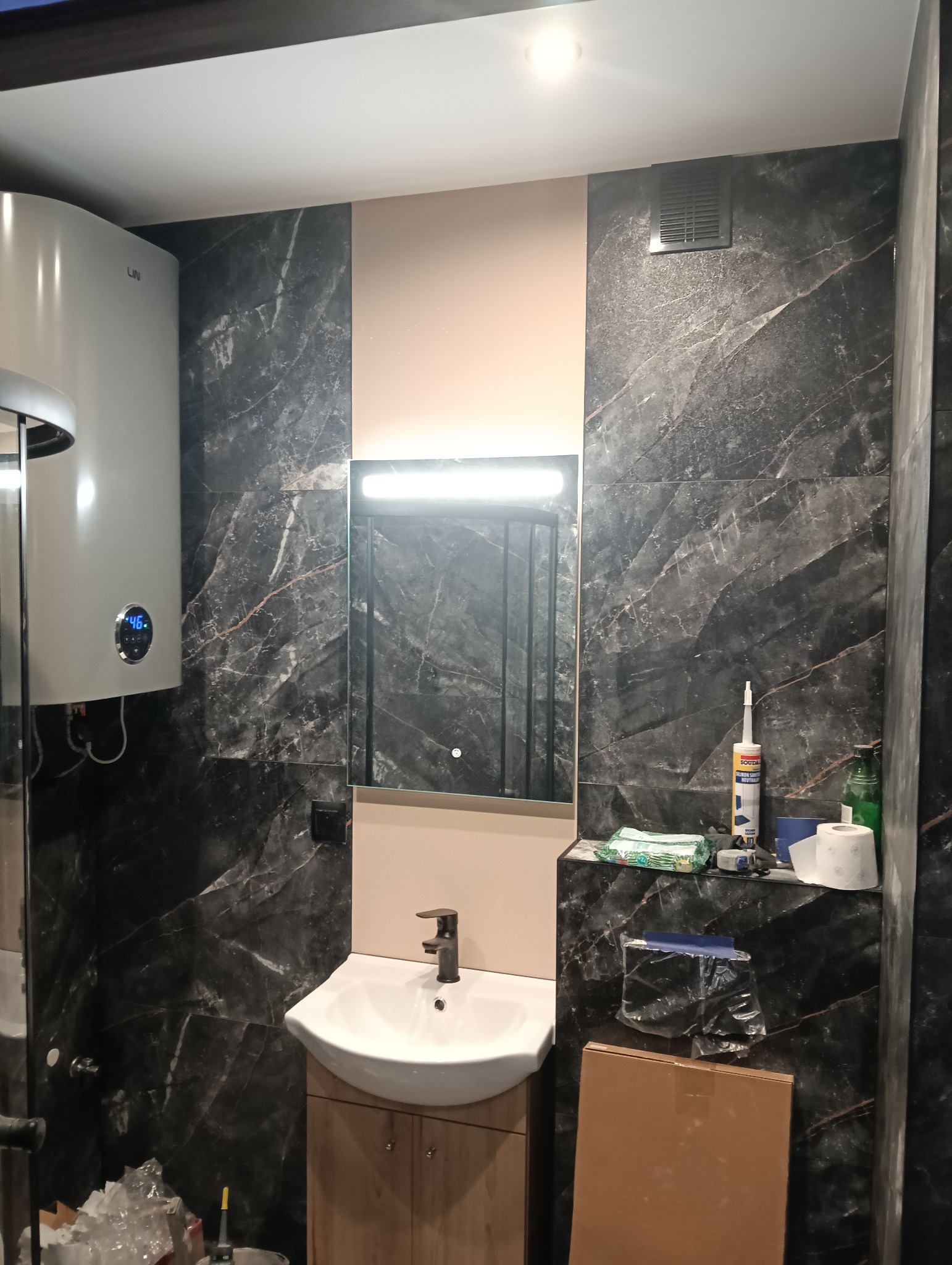 Wykończona łazienka z czarnymi płytkami imitującymi marmur, białą umywalką z szafką z jasnego drewna, podświetlanym lustrem i bojlerem po lewej stronie. Na blacie widoczne narzędzia i akcesoria.