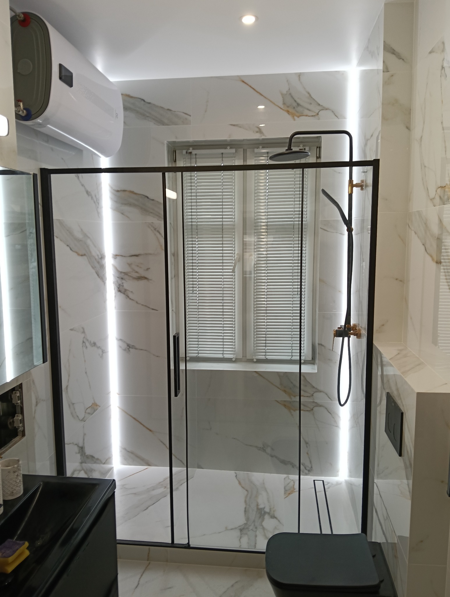 Nowoczesna łazienka z kabiną prysznicową typu walk-in z czarnymi ramami, marmurowe płytki z podświetleniem LED, wiszący bojler elektryczny i czarna armatura.