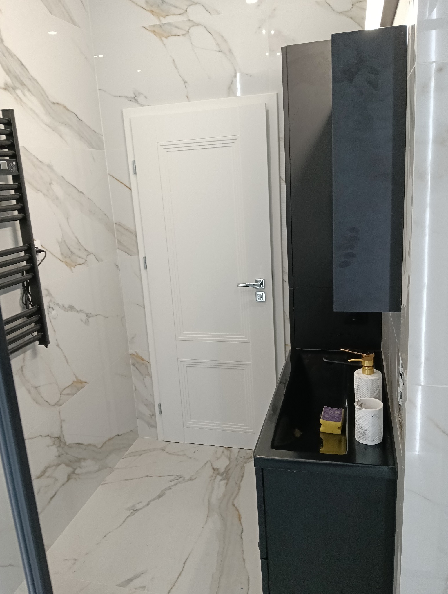 Nowoczesna łazienka z marmurowymi płytkami, czarną szafką umywalkową i białymi drzwiami, widoczny grzejnik drabinkowy.