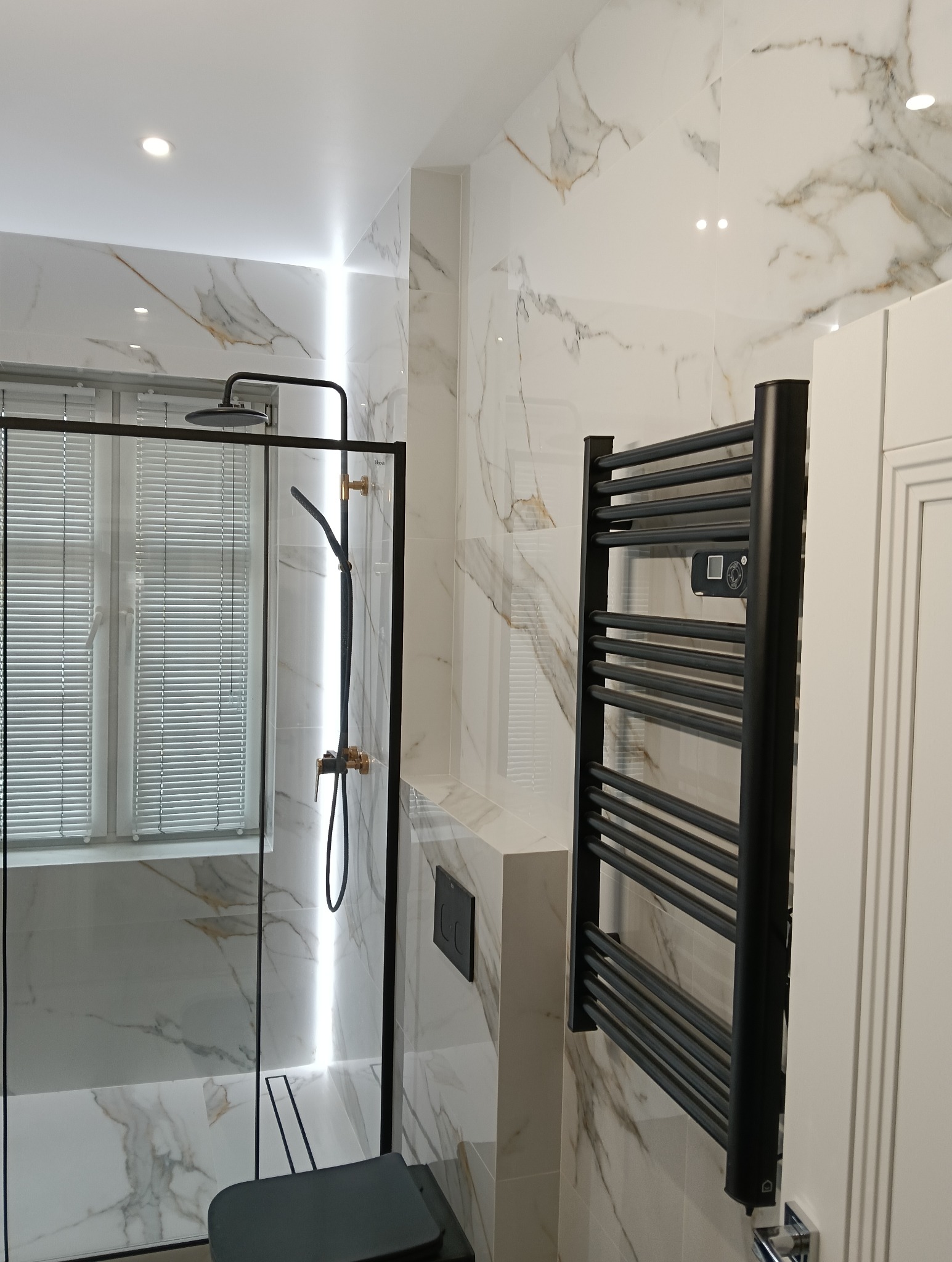 Nowoczesna łazienka z kabiną prysznicową typu walk-in, wykończona białymi płytkami imitującymi marmur, z czarną armaturą, grzejnikiem drabinkowym i minimalistycznym panelem WC.