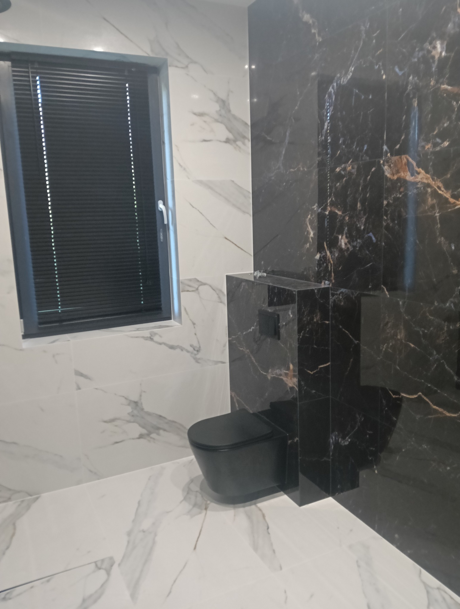 Nowoczesna łazienka z czarną wiszącą toaletą i marmurowymi płytkami w dwóch kolorach: białym i czarnym z złotymi żyłami, okno z czarnymi żaluzjami.