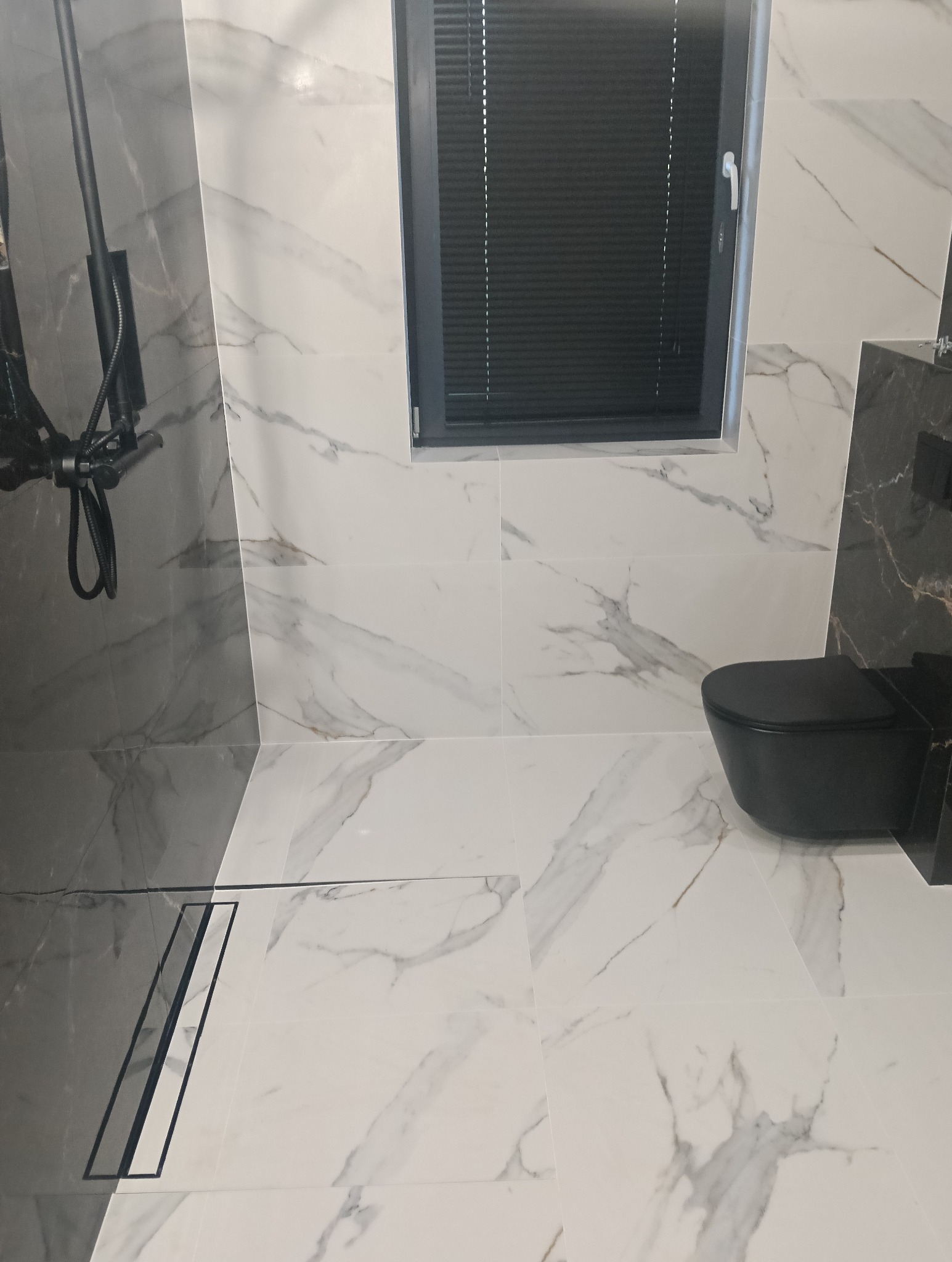 Nowoczesna łazienka wykończona białymi płytkami imitującymi marmur, z czarną armaturą, w tym deszczownicą i podwieszaną toaletą, oraz odpływem liniowym.