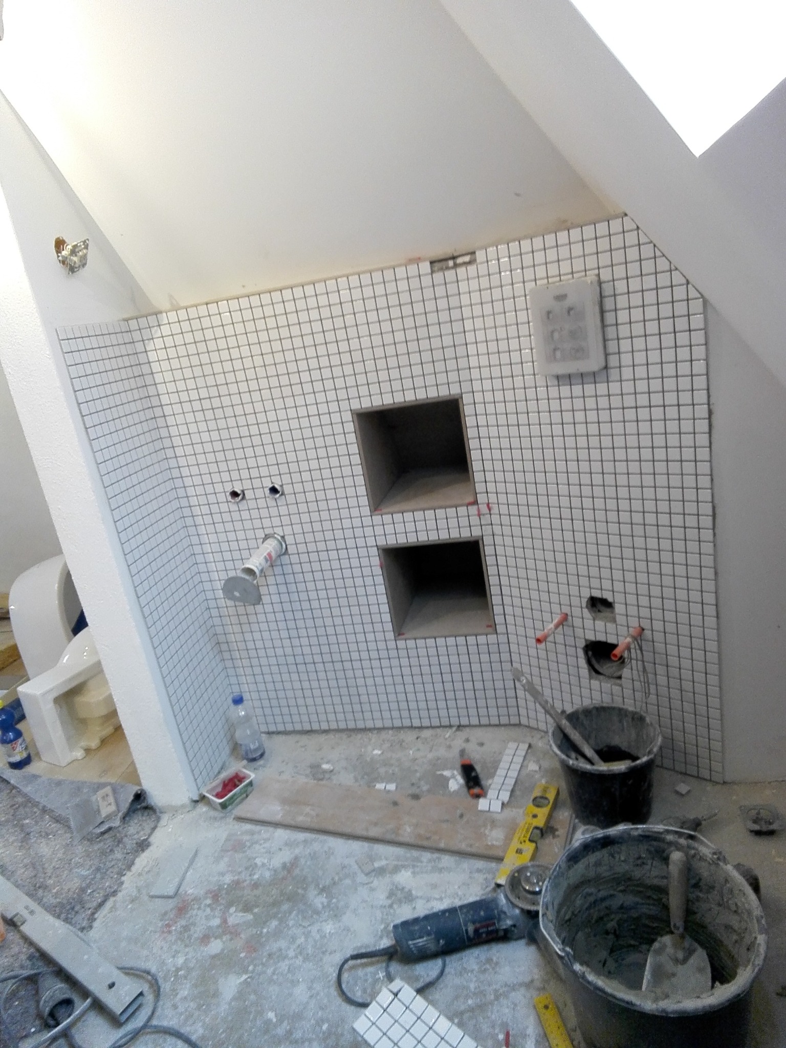 Łazienka w trakcie remontu: ściana wyłożona białą mozaiką z wnękami, widoczne narzędzia, wiaderka z zaprawą, szlifierka kątowa i fragment toalety w tle.