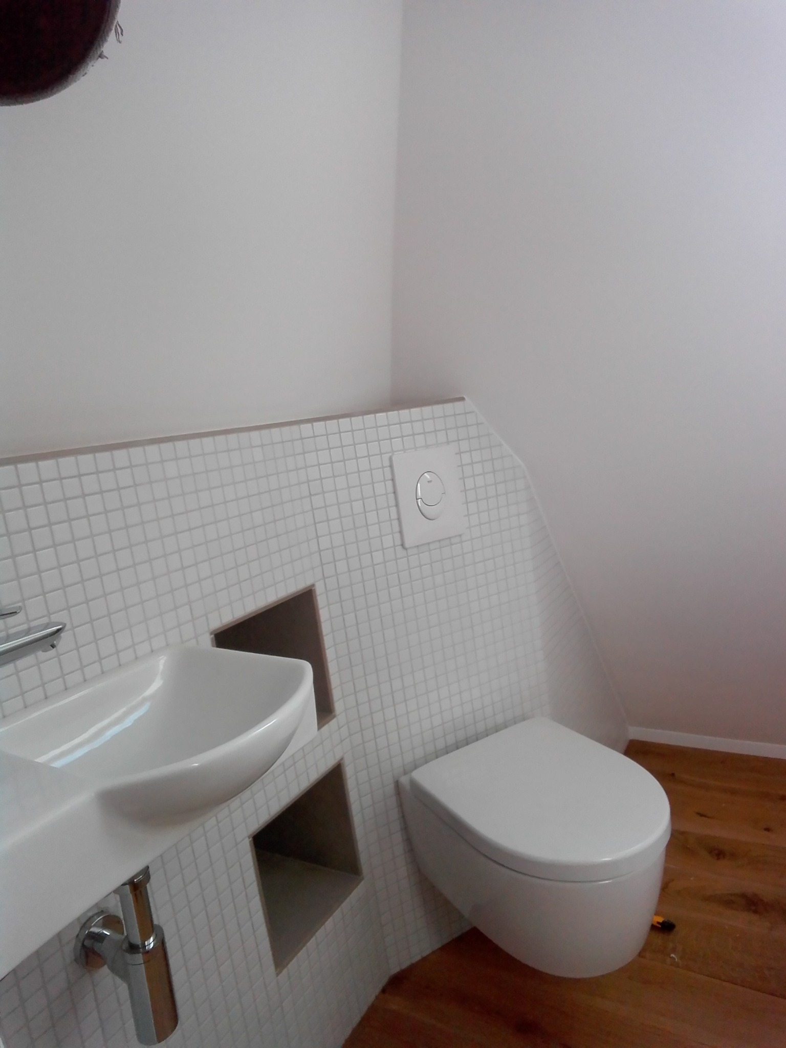 Nowoczesna łazienka na poddaszu z wiszącą miską WC i umywalką w zabudowie z białej mozaiki, drewniana podłoga.