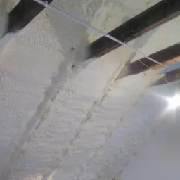 Poddasze z drewnianymi belkami po natrysku białej piany izolacyjnej, widoczne przewody elektryczne i fragment ściany.