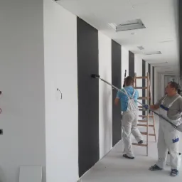 Dwóch malarzy w kombinezonach maluje ścianę w pionowe, czarno-białe pasy w przestronnym biurze, używając wałków na długich przedłużeniach; widoczna drabina i surowy stan wykończenia wnętrza.