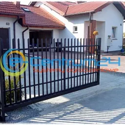 Ciemna, metalowa brama wjazdowa na posesję z domami o czerwonych dachach, widziana pod kątem, z logo firmy na pierwszym planie.