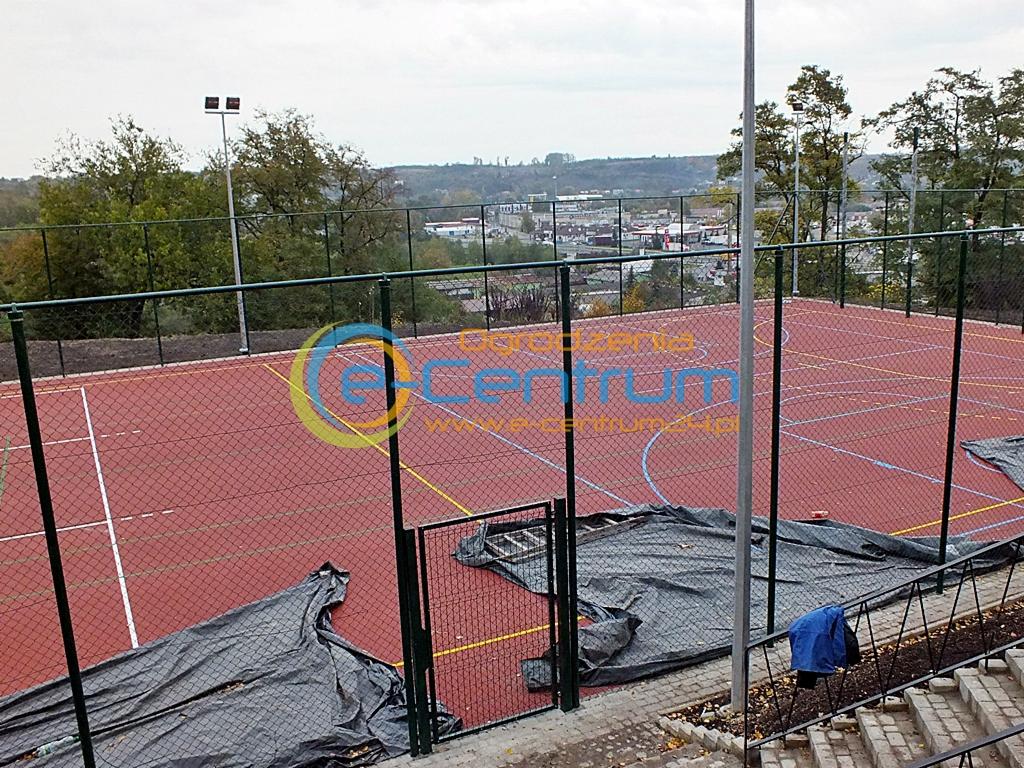 Boisko sportowe z zieloną siatką ogrodzeniową i otwartą furtką, widok z góry na tle zabudowań miejskich i drzew. Na boisku leżą plandeki.
