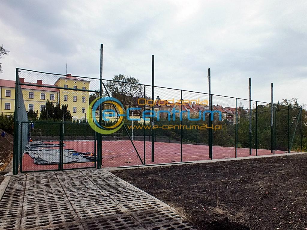 Siatka ogrodzeniowa okalająca czerwone boisko sportowe z zieloną bramą i słupkami na tle zabudowań miejskich w pochmurny dzień. Widoczny bruk i ziemia.