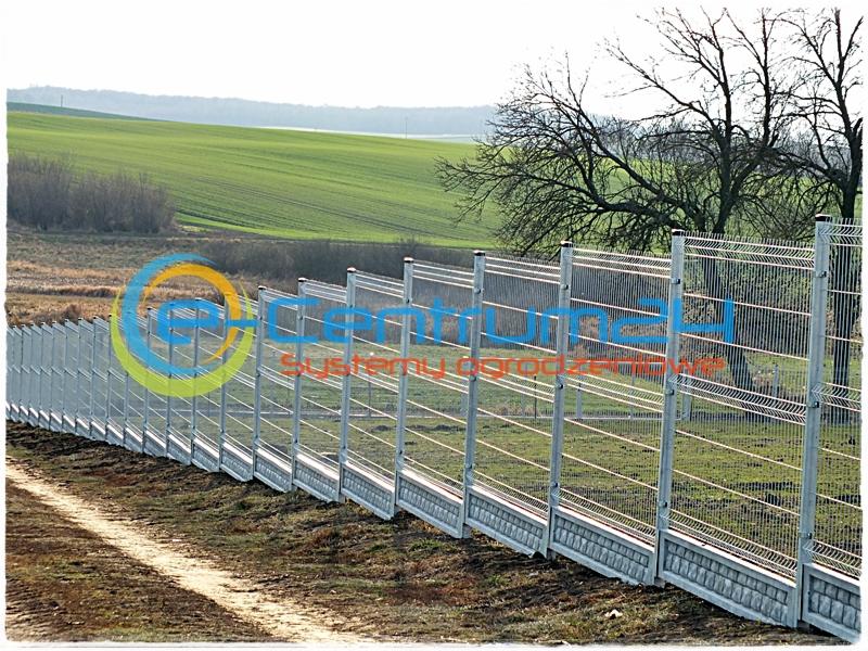 Długi, srebrny panel ogrodzeniowy z betonową podmurówką na tle zielonego pola i drzew. Widoczna polna droga wzdłuż ogrodzenia. Ujęcie z niskiej perspektywy.