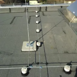 Instalacja odgromowa na dachu budynku z widocznymi elementami mocującymi i przewodami odprowadzającymi. Widok z góry na płaski dach z papy.