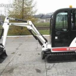Biało-czarna mini koparka Bobcat 323 na betonowej powierzchni, z widocznymi łyżkami i kabiną. W tle drzewa i budynek.