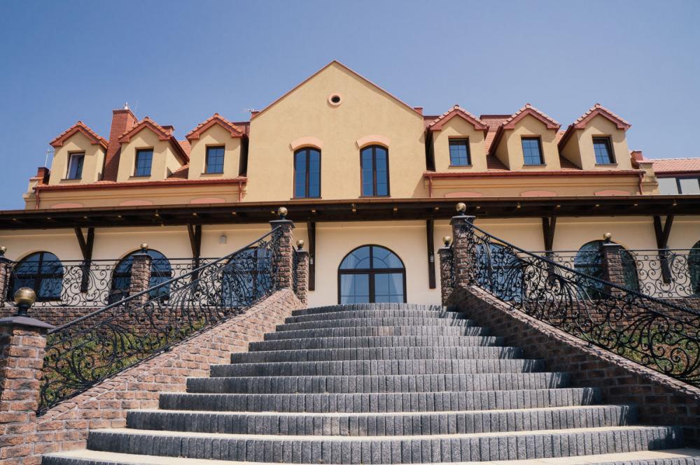 Elewacja budynku z beżową fasadą, schodami z szarej kostki i ozdobnymi, kutymi balustradami. Widok na budynek z dołu, w słoneczny dzień.