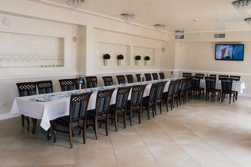 Sala bankietowa z długim stołem nakrytym białym obrusem i dekoracjami, gotowa na przyjęcie. W tle ściana z wnękami i telewizor.