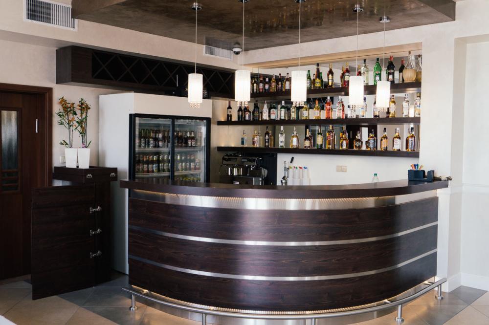 Elegancki bar z ciemnego drewna z metalowymi akcentami i oświetleniem punktowym. Półki wypełnione butelkami, lodówka z napojami.