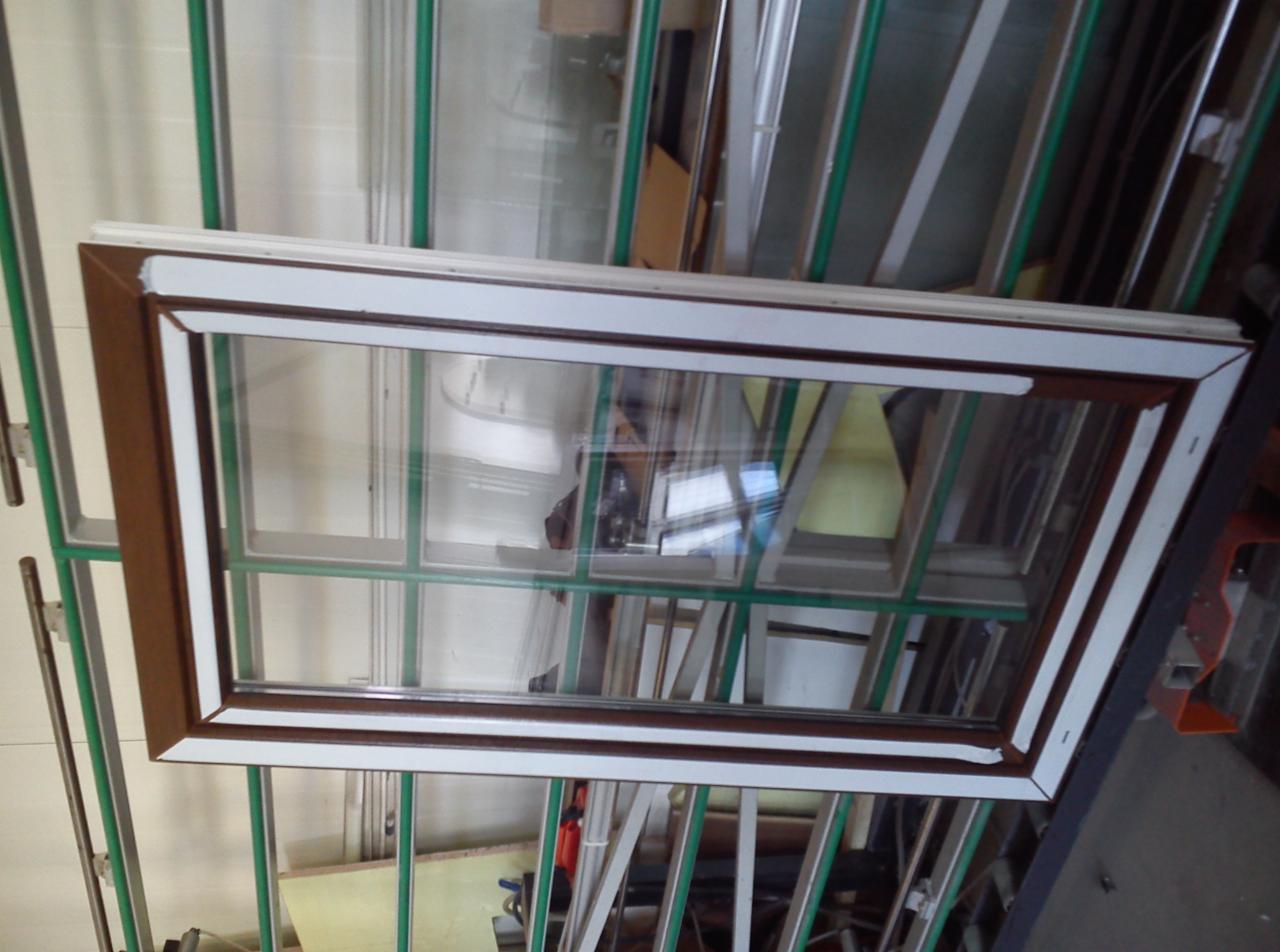 Biało-brązowe okno z szybami i szprosami, ustawione pionowo na konstrukcji z profili. Widoczne ślady montażu i otoczenie warsztatowe.