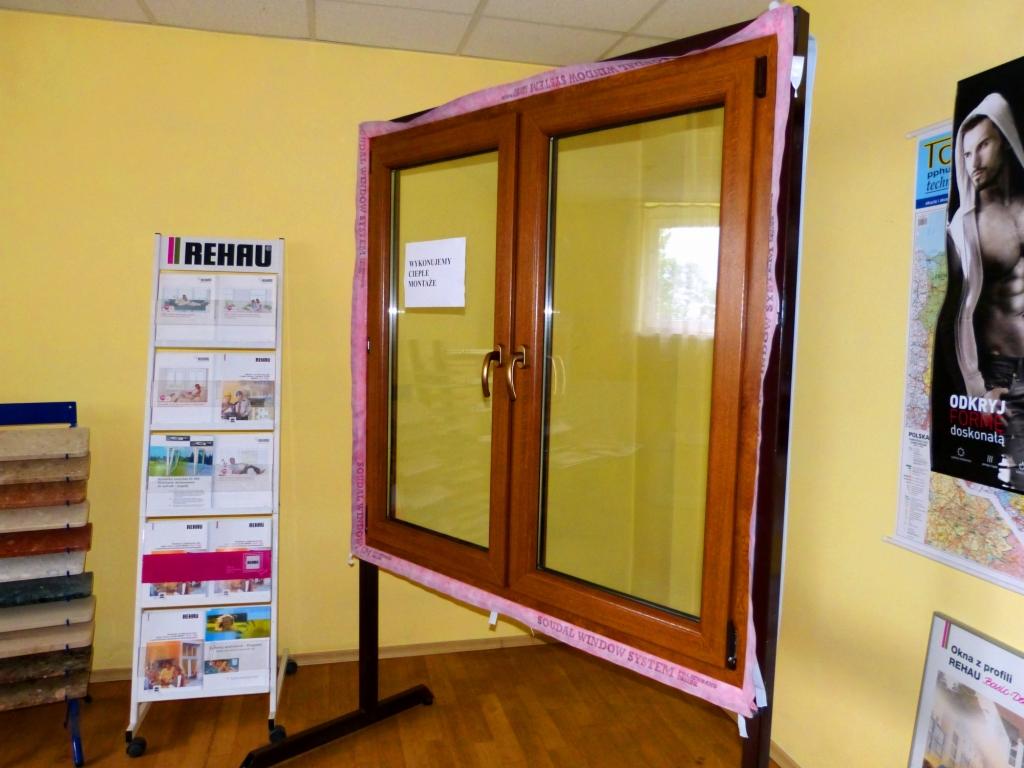 Prezentacja dwuskrzydłowego okna drewnianego z szybami, z ekspozytorem marki Rehau w tle, na tle żółtej ściany. Okno zabezpieczone różową pianką montażową.