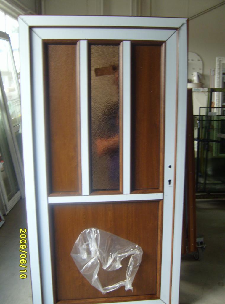 Brązowe drzwi z szybą i białą ramą, zabezpieczone folią. Widoczne elementy montażowe i data 2009/06/10. W tle inne drzwi i okna.