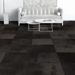 Płytki dywanowe w odcieniach szarości tworzące geometryczny wzór na podłodze, uzupełnione białym, pikowanym narożnikiem, okrągłymi stolikami i lampą łukową w minimalistycznym wnętrzu.