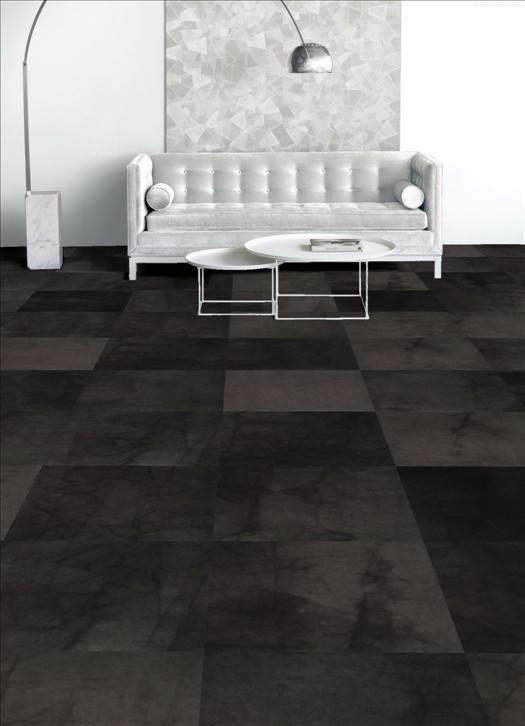 Płytki dywanowe w odcieniach szarości tworzące geometryczny wzór na podłodze, uzupełnione białym, pikowanym narożnikiem, okrągłymi stolikami i lampą łukową w minimalistycznym wnętrzu.