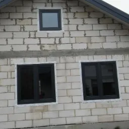 Surowy mur z betonu komórkowego z trzema zamontowanymi oknami z czarnymi ramami, zabezpieczonymi folią ochronną, widoczny fragment rynny i dachu.