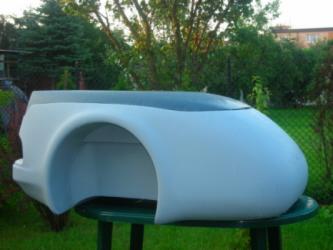 Element karoserii samochodowej z żywicy poliestrowej, prawdopodobnie błotnik lub fragment nadwozia, w kolorze szarym, umieszczony na zielonym stoliku ogrodowym na tle trawnika i zieleni.