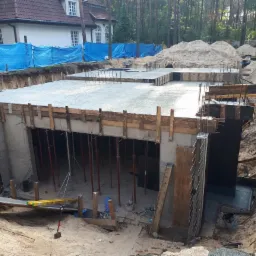 Wykop pod fundamenty budynku żelbetowego z widocznym szalunkiem, zbrojeniem i wylewką świeżego betonu. W tle dom jednorodzinny zabezpieczony niebieską plandeką.