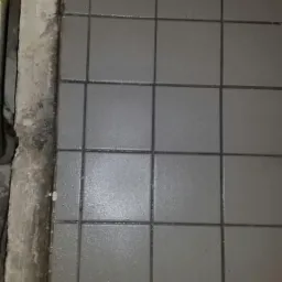 Ułożone szare płytki podłogowe z ciemną fugą, widoczne niedociągnięcia przy ścianie i ślady zaprawy.