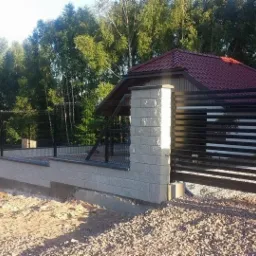 Nowoczesne ogrodzenie panelowe z podmurówką betonową i bramą przesuwną, na tle zieleni drzew i budynku z czerwoną dachówką. Widok z zewnątrz posesji, w słoneczny dzień.