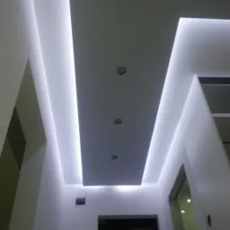 Podwieszany sufit z oświetleniem LED w nowoczesnym wnętrzu. Widoczne punkty świetlne i geometryczny kształt sufitu. Białe ściany, ciemne drzwi w tle.