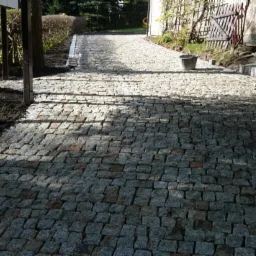 Ułożona kostka brukowa tworząca ścieżkę w ogrodzie, z widocznymi drzewami i rabatami po bokach. W tle budynek i elementy architektury krajobrazu.