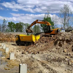 Żółta koparka JCB nabiera ziemię i wysypuje ją do żółtej ciężarówki na placu budowy z widocznymi betonowymi fundamentami i wykopem, w tle drzewa i błękitne niebo z chmurami.