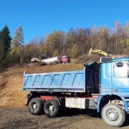 Niebieska wywrotka na tle wykarczowanego zbocza z pracującymi koparkami i wywrotką, błękitne niebo w tle.