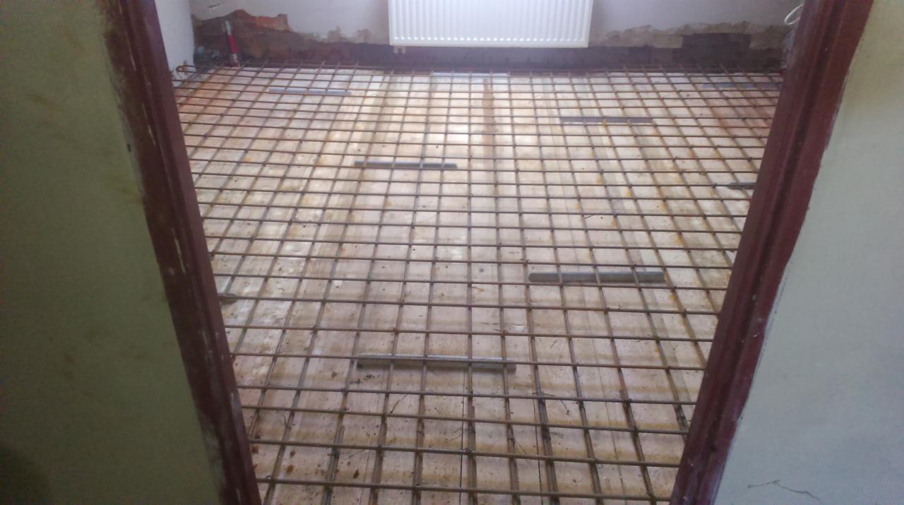 Zbrojenie pod wylewkę podłogową w pomieszczeniu w trakcie remontu, widoczne przez futrynę. Siatka zbrojeniowa ułożona na podłożu, grzejnik na ścianie w tle.