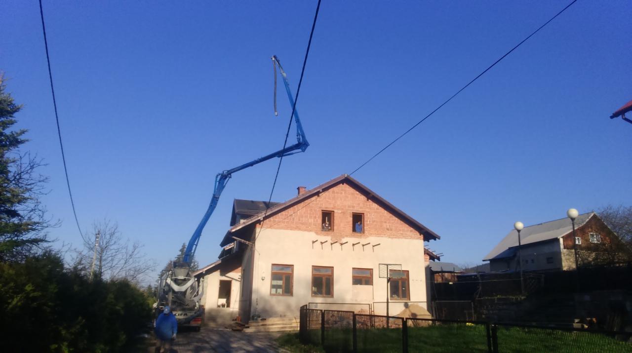 Dom w trakcie remontu z użyciem pompy do betonu, nieotynkowana elewacja, widoczne rusztowania i elementy konstrukcyjne. Błękitne niebo w tle.