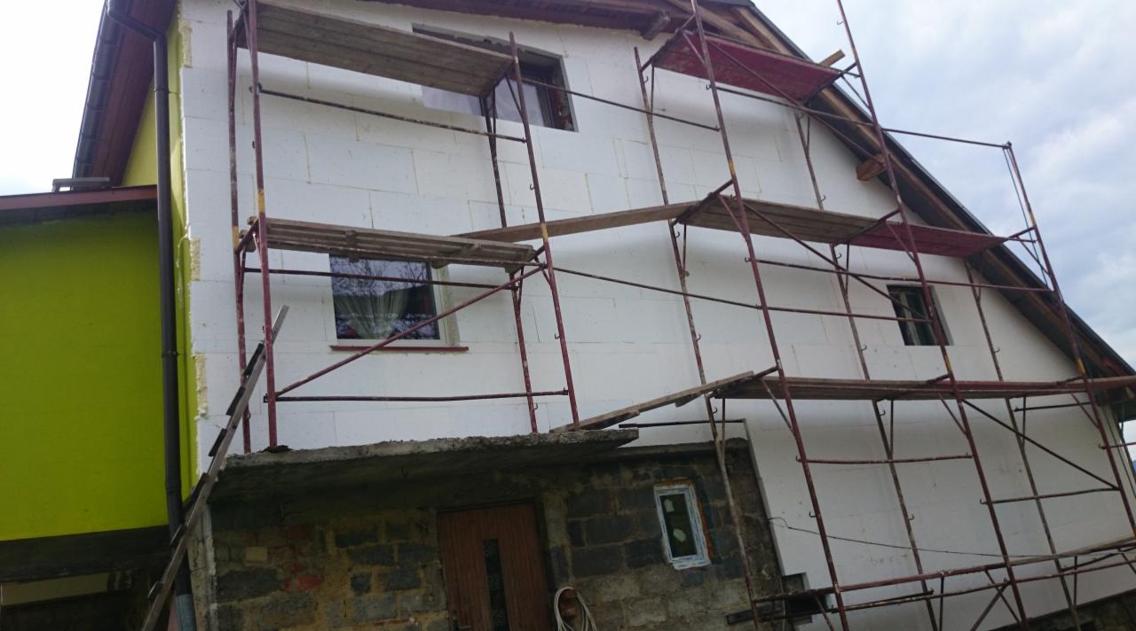 Dom w trakcie ocieplania styropianem, z widocznymi rusztowaniami i fragmentem zielonej ściany budynku sąsiadującego. Prace w toku, widok z dołu.