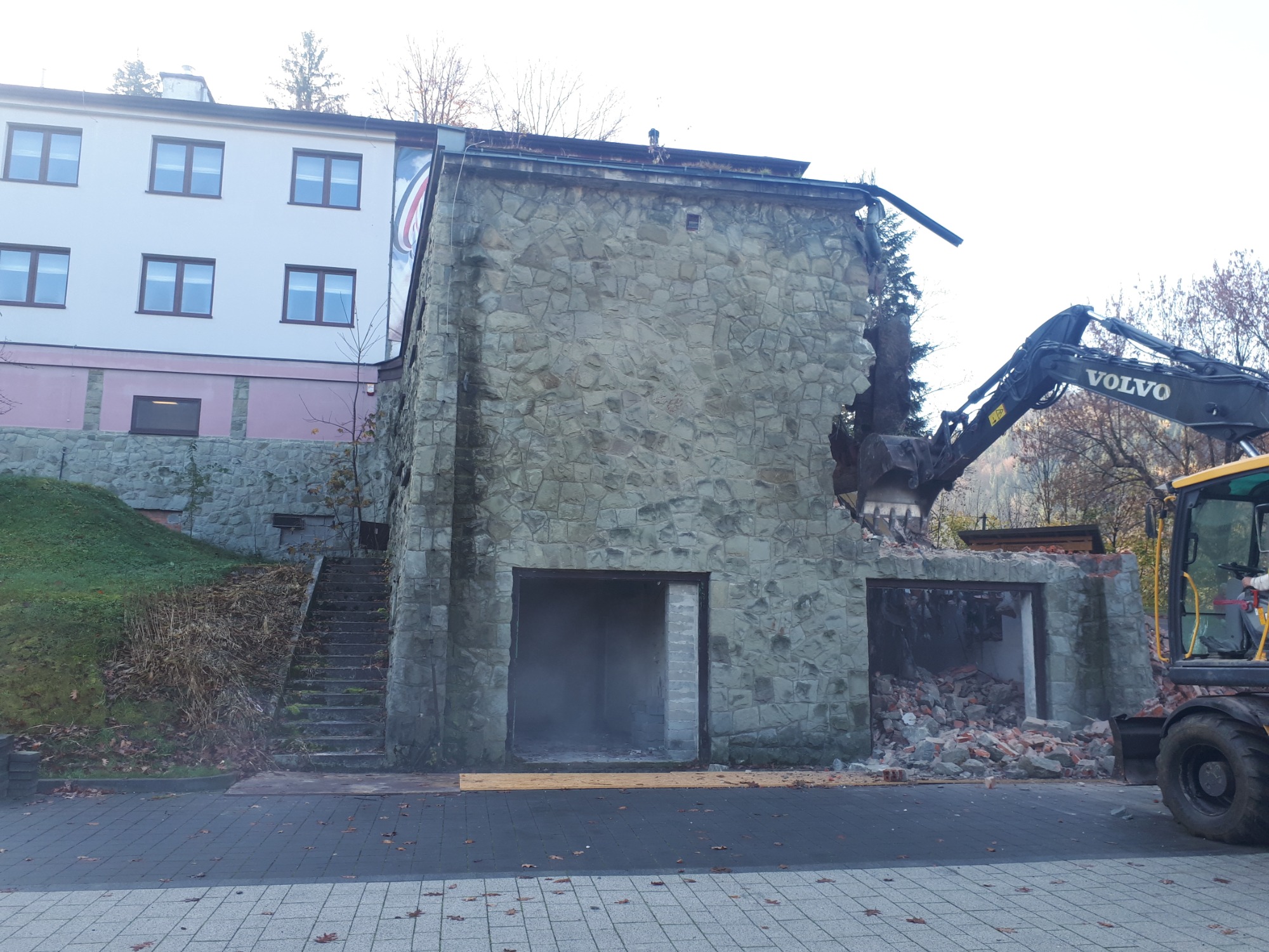 Częściowy rozbiórka kamiennego budynku z użyciem koparki Volvo, widoczne gruzy wewnątrz budynku, schody obok budynku porośnięte roślinnością.