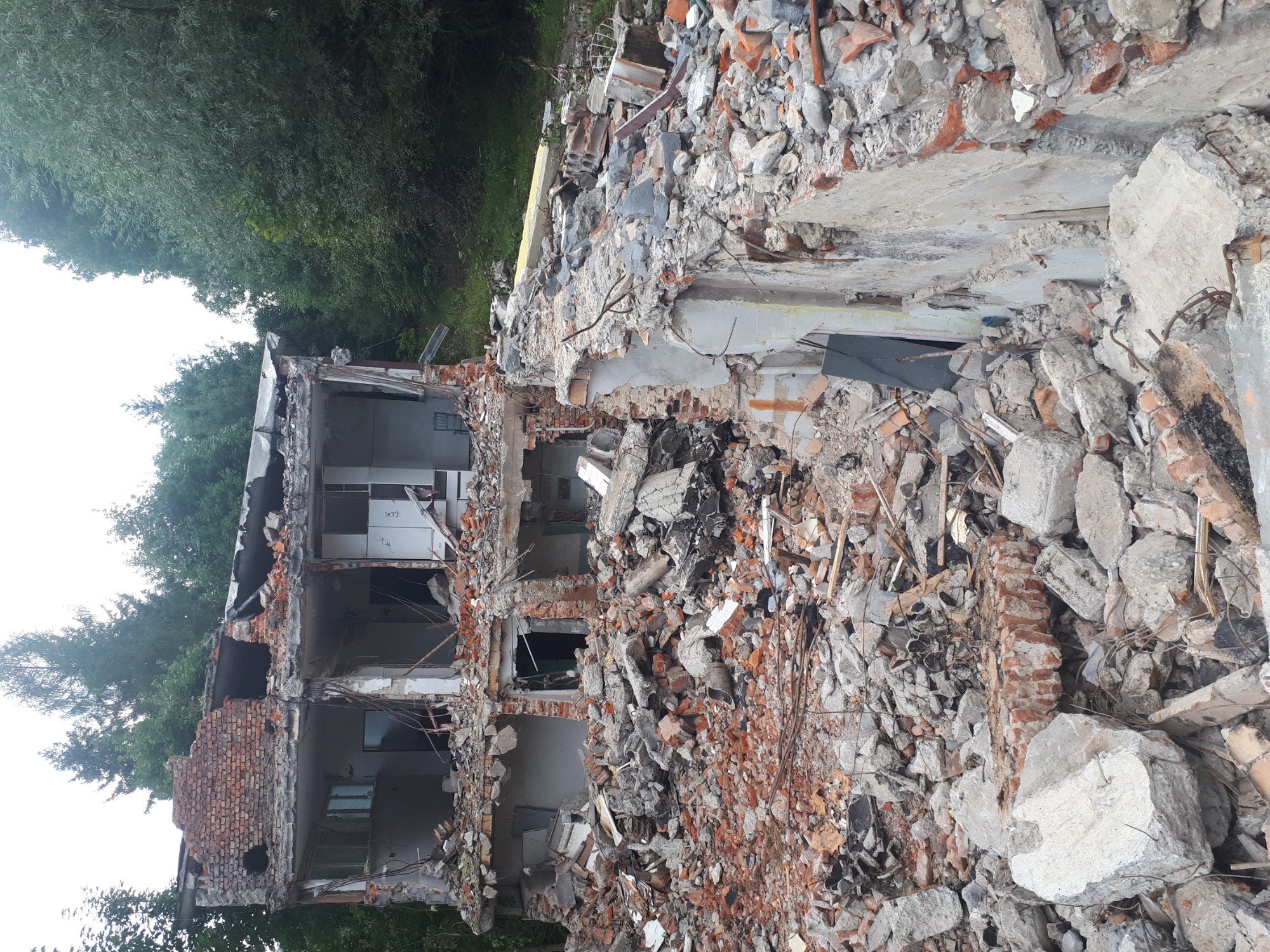 Zrujnowany budynek z odsłoniętymi fragmentami ścian, gruzem ceglanym i betonowym, otoczony zielenią drzew.