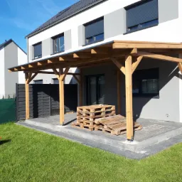 Nowoczesny dom z drewnianą konstrukcją zadaszenia tarasu na betonowej kostce, stos palet na tarasie, zwinięty wąż ogrodowy na trawniku.