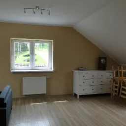 Beżowy pokój na poddaszu z drewnianą podłogą, białymi meblami, oknem z widokiem na zieleń i lampą sufitową na listwie. Ustawienie mebli i przestronność.